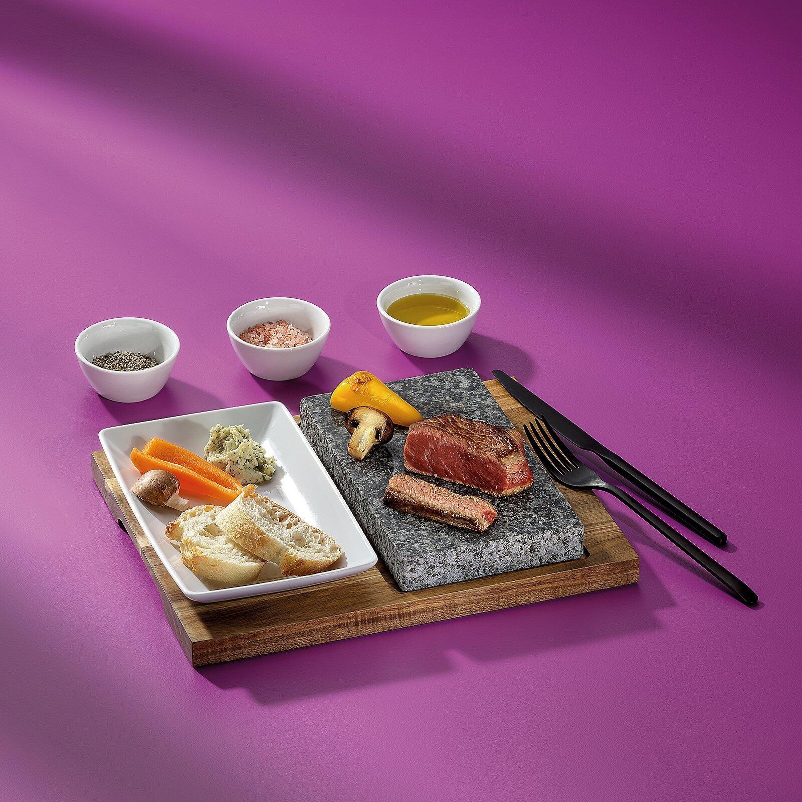 Servierbrett "Heißer, Country" mit Steak, gegrilltem Gemüse, Schälchen mit Gewürzen und Messern auf hellem Holzuntersetzer.
