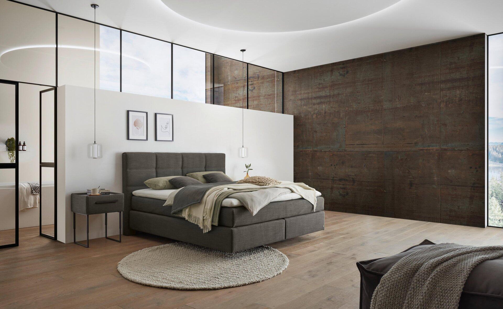 Boxspringbett Luxor Line BX mit grauem Kopfteil, Rechtecksteppung und Schwebeoptik-Füßen, eingebettet in ein Schlafzimmer mit Dielenparkett und Nachttisch.