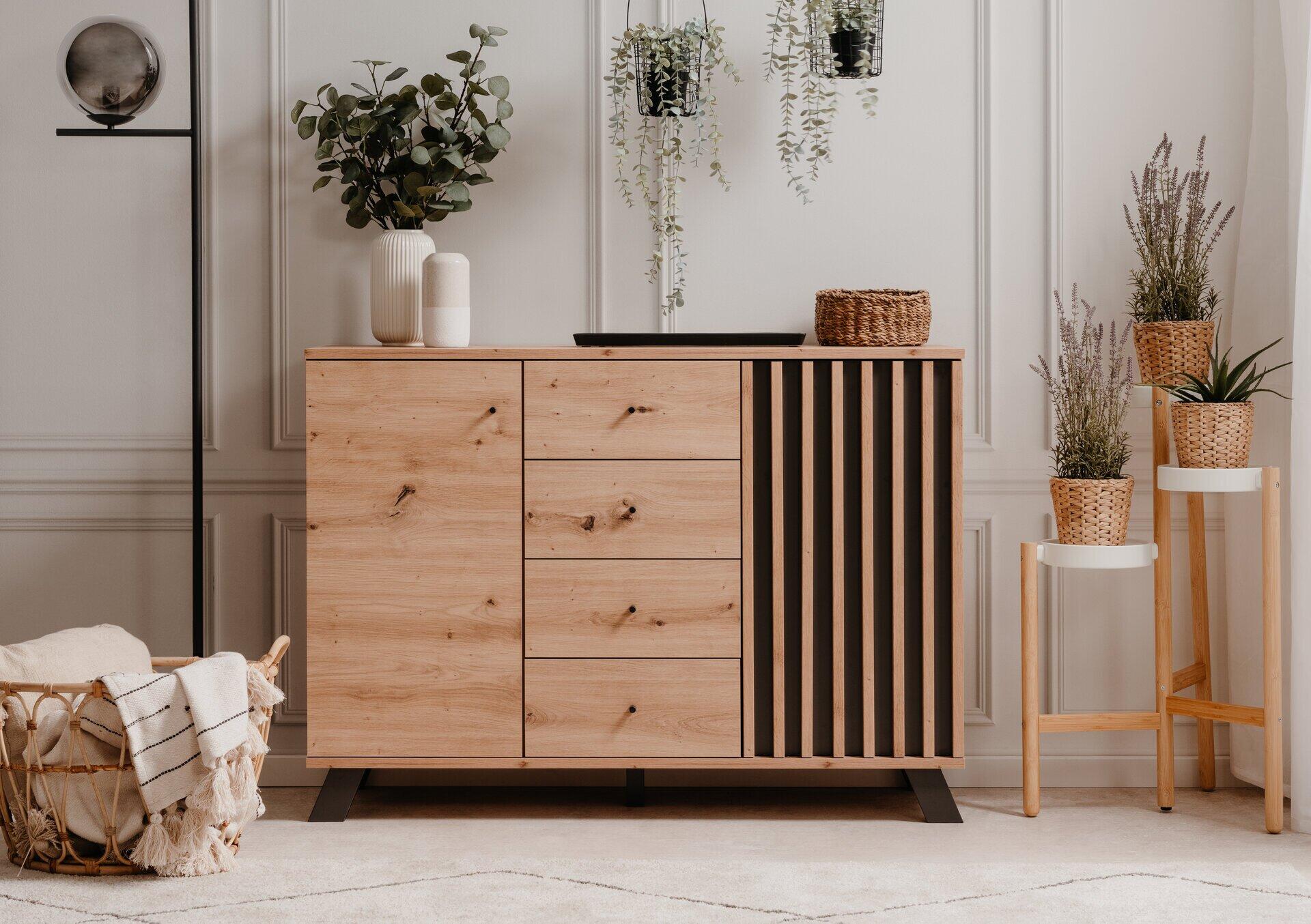 Moderne Kommode Medan 01B mit vier Schubladen, zwei Türen, schwarzen Füßen und schwarzen Griffen, präsentiert in einem stilvollen Wohnzimmer mit angrenzenden dekorativen Elementen.