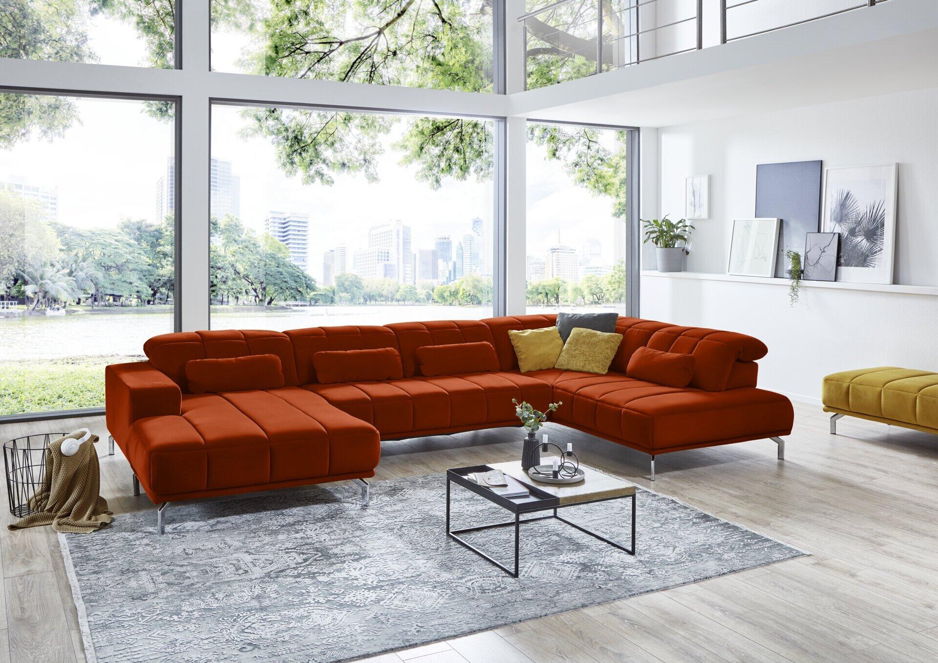 Orangefarbenes Ecksofa Modell Klagenfurt mit Samtbezug, kastenförmiger Quiltung der Rückenlehnen und verstellbaren Kopfteilen auf silberfarbenen Füßen.
