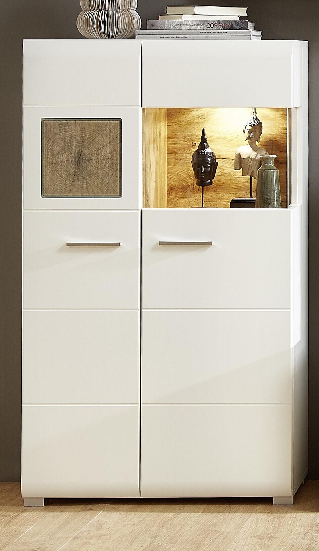 Vitrine Fun Plus mit weißen, glänzenden Türen, Milchglasfenster und integrierter LED-Beleuchtung, die dekorative Elemente auf einer dahinterliegenden Ablagefläche beleuchtet.