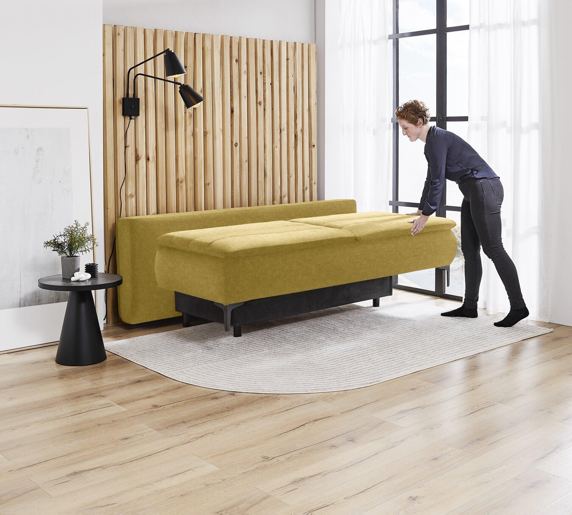 Boxspringsofa Melly im geöffneten Zustand, zeigt die ausziehbare Liegefläche mit den Maßen 140x200cm, neben einem Beistelltisch und einer Frau.
