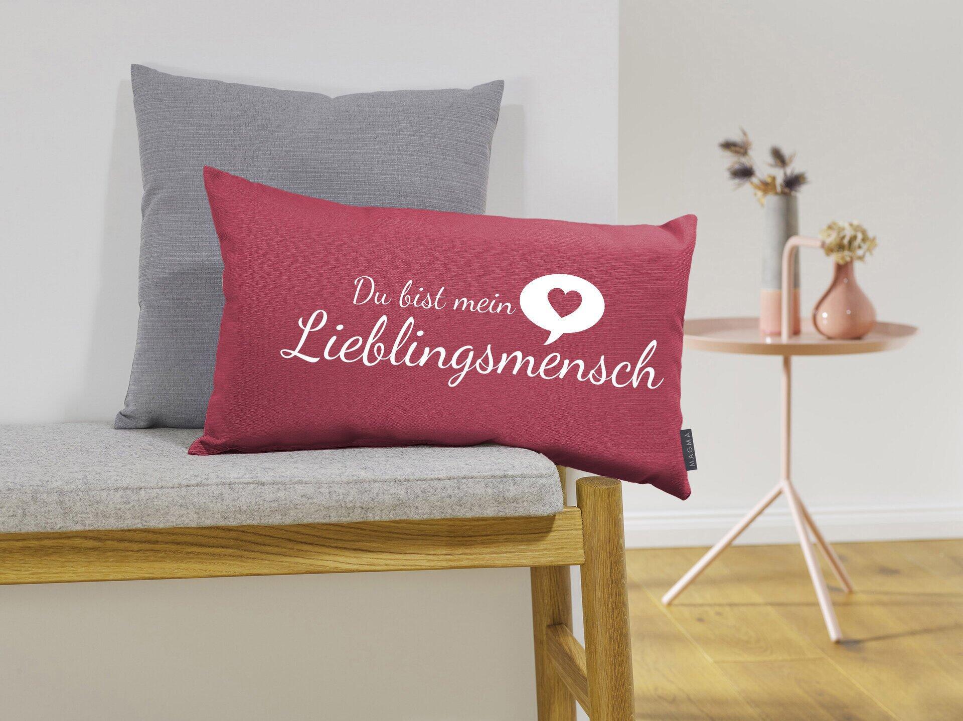 Dekokissen "Lieblingsmensch" mit tiefrotem Stoff, weißem Schriftzug und silbrigem Rand auf einer weißen Bank.