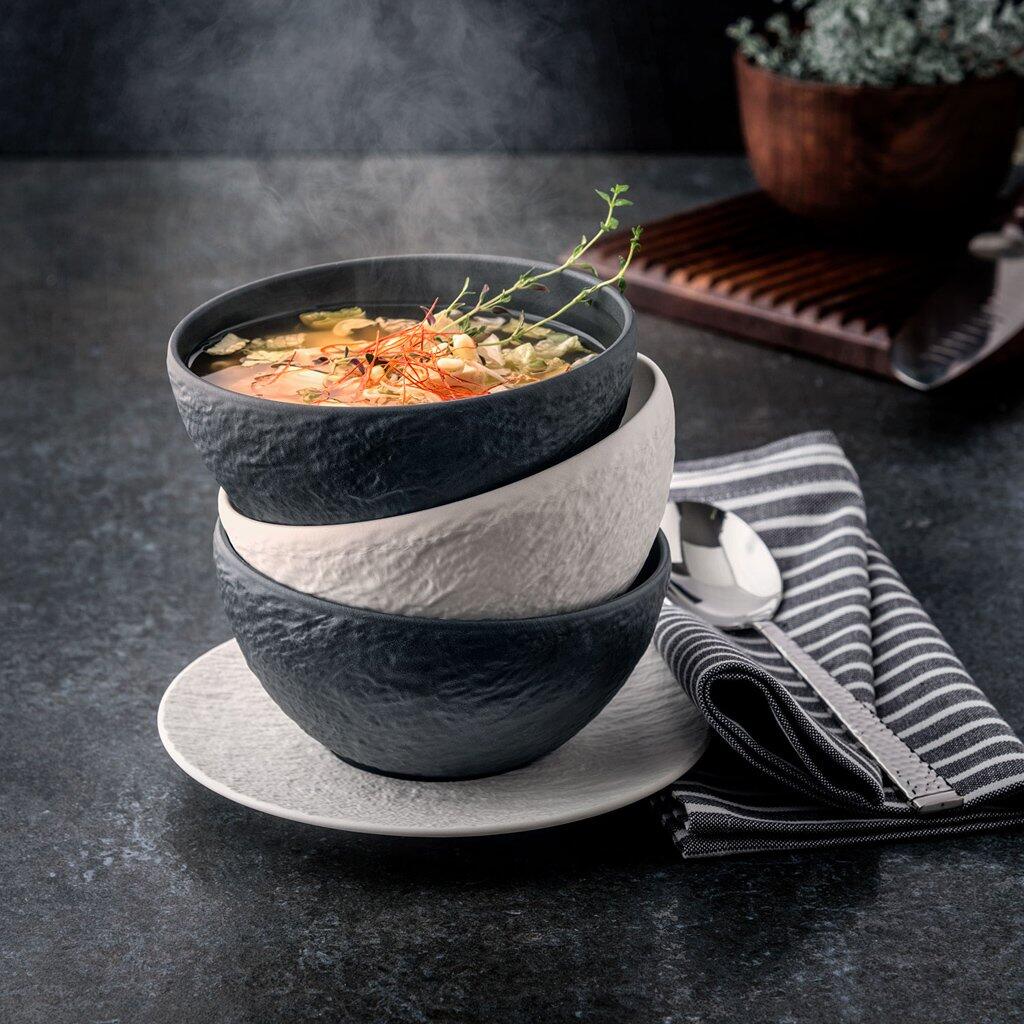 Gestapelte dekorative Schalen der Serie Manufacture Rock von Villeroy & Boch mit dunklen Farbnuancen, eine davon mit einer dampfenden Suppe auf einem Teller, aufgenommen aus Vogelperspektive.